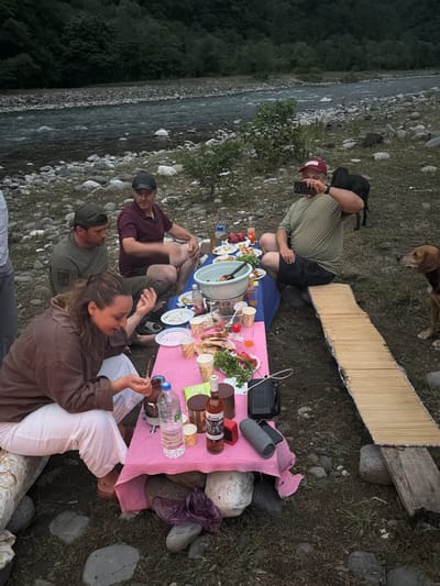 The image depicts a group of people having a picnic near a river in Wazabaa, Abkhazia. Four individuals are seated around a low table covered with a pink tablecloth, laden with various food items and drinks. A woman in a brown hoodie and white pants is in the foreground, holding a bottle and looking at her hands. To her right, a man in a green checkered shirt is taking a picture with his phone, with a black dog resting on his shoulder. Two other men are seated further back; one in a maroon shirt is looking towards the table, and another in a green shirt is also near the table. Two dogs are present in the scene, one black with the man and a brown one to the right, observing the gathering. The environment is natural, with a river flowing in the background, lined by a rocky shore and dense vegetation on the opposite bank. The ground is covered with grass and stones, and a makeshift seating area with a woven mat is visible on the right.