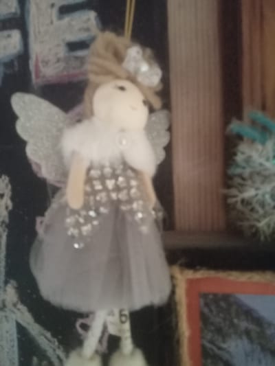 A blurry, close-up, vertical shot features a Christmas ornament of an angel. The angel has light-colored yarn hair and a white fluffy collar. Its dress is light gray with silver embellishments. The angel has white, glittery wings and appears to be hanging from a string. The background is dark and out of focus, with hints of a blackboard or chalk drawings, and a blurred wooden surface. A framed picture is partially visible on the right side of the image, and a blurry evergreen branch with artificial snow is also present.