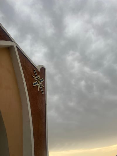 The image shows a close-up of a building's architectural detail against a cloudy sky. The building features an arched entrance with a textured reddish-brown surface and a cream-colored trim. A decorative emblem, resembling a star or a stylized cross with blue and green accents, is affixed to the reddish-brown facade. The sky is overcast with heavy, gray clouds, and a faint sliver of yellow light is visible at the bottom of the frame, suggesting it might be dawn or dusk. The location of Wai A, Nigeria is not identifiable from the visual information presented in the image.