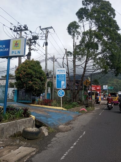 The image depicts a roadside scene in Indonesia, specifically near a PLN (Perusahaan Listrik Negara) electricity facility and an electric vehicle charging station, as indicated by prominent signage. The sky is overcast.
A two-lane asphalt road, partially visible on the right with a white dashed line, carries multiple motorbikes and a yellow minivan/taxi traveling away from the viewer. One motorcyclist in a red jacket and helmet is prominent.
To the left of the road, a concrete curb abuts a blue-painted concrete ramp leading into a fenced compound with a blue metal gate. A discarded tire rests on the ground near the curb. Two primary signs are visible: a large rectangular white and blue sign on a blue pole displaying "PLN PACAMACAN" with the PLN logo, and a smaller blue rectangular sign below it stating "SPKLU PLN ELECTRIC VEHICLE CHARGING STATION" with an EV charger icon and a blue directional arrow pointing left.
Multiple utility poles with a dense network of black electrical wires and transformers are situated above the compound. Lush green vegetation, including a large rounded bush and several tall evergreen trees (some with bare branches), frames the left and central portions of the image. A yellow diamond-shaped road sign indicating an intersection is visible further down the road on the right, along with various commercial banners. Green mountains are discernible in the distant background under the cloudy sky.
