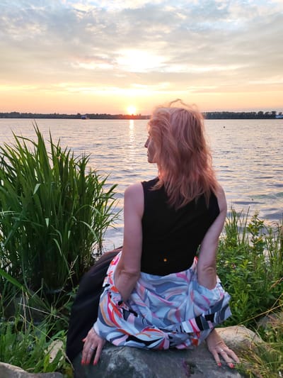 A woman with pink hair is sitting on a rock by the water, facing away from the camera. She is wearing a black top and a skirt with a colorful abstract pattern. The sun is setting over the water, casting a warm glow on the scene. The water is calm, with gentle ripples. There is tall grass and some smaller plants growing around the rocks. In the distance, there is a treeline and a small boat on the water. The sky is filled with soft clouds, painted in shades of orange, pink, and blue. The location is the shore of a body of water in Chelganovo, Russia.