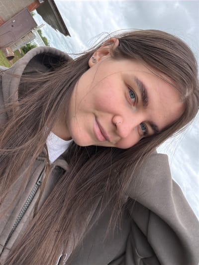 A young woman with long, brown hair and blue eyes is taking a selfie outdoors. She is wearing a grey hooded sweatshirt and a white t-shirt. She has a nose ring and earrings. The background shows a cloudy sky and a building with a metal roof. The image is shot from a close-up angle, with the woman's face filling most of the frame. The location is not specified as being in the city of Ромодан, Russia.