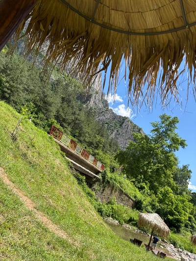 The image captures a scenic view of a natural environment, possibly a park or a riverbank, with a mountainous backdrop. In the foreground, a thatched umbrella provides shade, with two empty lounge chairs positioned on a grassy area next to a body of water. Further back, a small bridge with red and white railings spans across a slope covered in lush green grass and vegetation. The bridge is situated on the side of a steep incline, leading up towards a rocky mountain face. Dense green trees and foliage cover the slopes and mountainside, with a clear blue sky dotted with white clouds overhead. The overall impression is one of a tranquil outdoor setting, likely a popular spot for recreation and relaxation. The location is identified as Zgorigrad, Bulgaria, suggesting it's a natural landmark or recreational area within or near the city.