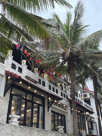 A multi-story building, likely a commercial establishment, stands prominently in An Hải Ward, Vietnam, partially obscured by two tall palm trees with green fronds. The building features white-painted brickwork or stone cladding on its exterior, with a textured base of small, light-colored pebbles. Red-tiled awnings are visible over ground-floor windows and possibly balconies. The ground floor displays large, dark-framed glass doors and windows, one of which is open, reflecting interior lights. A string of exposed bulb lights hangs beneath the lower awning. Upper levels feature multiple balconies with white balustrades and decorative yellow/gold square lantern-like elements on pillars. Numerous red spherical lanterns are suspended from tree branches and overhead structures on the balconies, with a few multicolored lanterns visible on a higher terrace. Several outdoor air conditioning units are mounted on the white walls of the building's upper floors. The sky is overcast. No people or specific actions are visible.