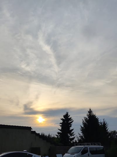 This image captures an outdoor scene at dusk in Czapury, Poland. The sky is filled with a mixture of clouds, with the setting sun peeking through, casting a warm, golden glow. The sun's rays create a dramatic effect as they are diffused by the atmospheric conditions.
In the foreground, a wall of a building is visible, likely a house or a garage, with a tiled roof. In front of the wall, parts of two vehicles are present. The rear of a white van with a roof rack is prominently displayed, suggesting it is parked. Next to it, the front of a white car is partially visible, with some of its features obscured.
Behind the wall and vehicles, a line of evergreen trees, likely fir or pine trees, stands silhouetted against the sky. Some of these trees are quite tall, reaching towards the upper half of the frame. Electrical wires are strung across the scene, indicating the presence of infrastructure.
The overall mood of the image is tranquil and serene, characteristic of a late evening. The soft light and the silhouette of the trees contribute to a peaceful atmosphere. The presence of vehicles suggests a residential or possibly a business setting. No people are visible in the image. No text is discernable.
