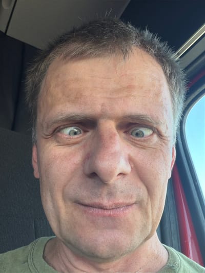 The image is a close-up, selfie-style photograph of a middle-aged Caucasian man with light-colored eyes and fair skin, taken outdoors during the day. He is wearing a green t-shirt. His hair is short and appears to be a mix of brown and gray. The man is looking directly at the camera with his eyes crossed. The background shows a slightly out-of-focus interior, possibly of a vehicle, with a dark textured surface on the left and a red object on the right, consistent with a car door or frame. A bright blue sky is visible through a window on the right side of the frame. The image was taken in North Platte, United States of America.