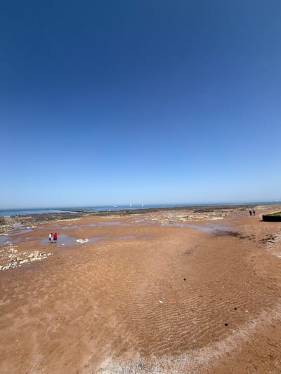 The image shows a beach scene in the city of Thanet, United Kingdom. The view is split between a clear blue sky occupying the left half of the frame and the sandy beach on the right. Several small boats can be seen far out on the ocean. A few people are visible near the bottom of the frame, standing close to the water's edge on the wet sand and rocks. The sandy beach shows ripple patterns, indicating it is low tide and the water has receded. Further up the beach, the sand is darker and more textured.