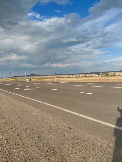 A photograph captures an asphalt road with white lane markings, stretching into the distance under a partly cloudy sky. The sky is a mix of blues and grays, with cumulus clouds drifting overhead. To the left of the road, a gravel shoulder is visible, and beyond that, a flat, arid landscape with sparse vegetation and distant, low hills. Several lampposts line the roadside. On the left side of the road, a yellow bus stop shelter with a blue roof is present. In the far distance, a collection of low-rise buildings can be seen. A shadow of a person with a raised arm and hand is cast on the right side of the road. The scene appears to be in a rural or semi-rural area, possibly on the outskirts of a settlement. The location is identified as Қапанбұлақ ауылдық округі, Kazakhstan.