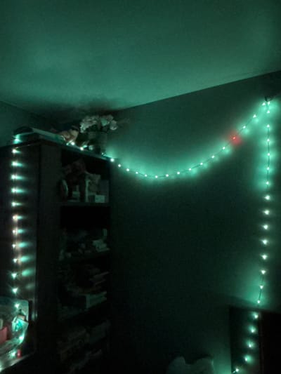 The image depicts an indoor scene, likely a bedroom, illuminated by string lights. The dominant color is a deep green, cast by the lights themselves and possibly the surrounding environment. On the left side of the frame, a dark bookshelf is visible, its shelves partially obscured by darkness and the glow of vertical strings of white and teal lights. On top of the bookshelf, a bouquet of light-colored flowers is arranged in a vase, with some objects beneath the flowers. A string of green lights trails across the upper wall and then down the right-hand wall, with one red light interspersed within the green ones. The overall impression is one of a dimly lit, possibly decorated, room. There are no individuals present in the image. The location of Novaya Usman', Russia, is not discernible from the visual information provided in the image.