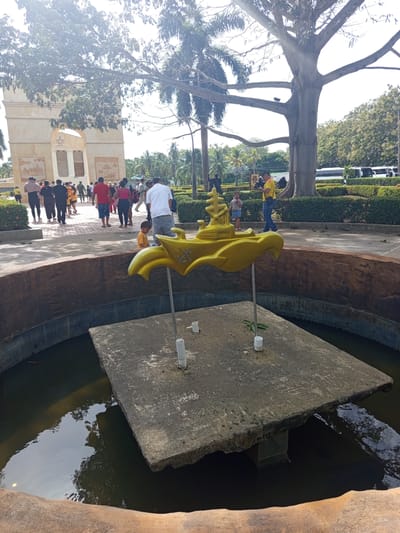 In Coveñas, Colombia, a group of people are gathered near a large stone archway. In the foreground, a yellow, genie lamp-shaped sculpture is positioned on a concrete platform within a water feature. The sculpture has anchor motifs. The environment is sunny with lush green trees and foliage surrounding the area. A large, old tree with thick branches dominates the background. Several buses are parked in the distance.
