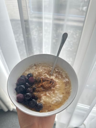 The image depicts a close-up, high-angle view of a white ceramic bowl filled with a creamy, light-colored porridge, likely oatmeal, held in a human left hand. The porridge is topped with a cluster of dark blue berries, some exhibiting a frosted appearance, indicating they are frozen. Also visible are a dollop of light brown peanut butter, a drizzle of translucent amber liquid (likely honey or syrup), and a dusting of reddish-brown spice, possibly cinnamon, covering the surface. A silver-colored metal spoon is inserted into the porridge, angled towards the top right. The background consists of a window covered by two sheer white curtains, through which an indistinct outdoor, naturally lit environment is partially discernible. The scene is located in Wanneperveen, Netherlands.