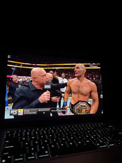 Here is a detailed description of the image:

The image shows a close-up view of a laptop screen displaying a UFC (Ultimate Fighting Championship) broadcast featuring Sean Strickland, a mixed martial artist. Strickland, shirtless and wearing the championship belt, is visible in the frame. Next to him is an interviewer. The screen also shows text and logos, including "UFC 328" and the Paramount+ logo. The laptop keyboard is visible on the right side of the frame.

There is no indication in the picture that the image or subjects are physically present in the city of Sant Vicent del Raspeig, Spain. The image is purely of the laptop screen.