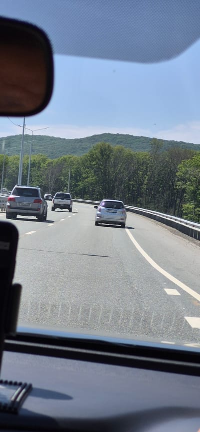 The image is taken from inside a vehicle, looking forward through the windshield, with a rearview mirror visible in the top left corner. The vehicle is on a highway with multiple lanes, indicated by white lane markings. The road is paved and appears to be in good condition.

In front of the vehicle, there are several other cars on the highway. The closest car ahead is a silver hatchback. To the left of this car, there is a white pickup truck, and further to the left and slightly ahead is a silver SUV. The license plate of the SUV is visible and appears to be in Cyrillic script.

The highway is flanked by a metal guardrail on the right side. Beyond the guardrail, there is a dense forest with green trees, indicating a rural or semi-rural setting. In the background, rolling hills covered in trees form a backdrop under a clear blue sky with scattered clouds.

The scene suggests a daytime journey on a highway surrounded by nature. There are no visible people other than the presumed driver and passengers of the vehicles. The presence of Cyrillic script on the license plate suggests the location is in a Russian-speaking region. Given the context provided about Gorodechnoe, Russia, it is highly probable this scene is located there, though the image itself does not contain any explicit identifiers of this specific location.