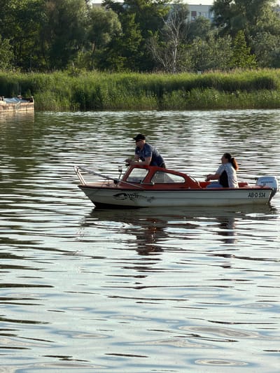 The image depicts two people in a small motorboat on a body of water, likely a lake or river, near a grassy bank with trees. The boat is white with red trim and has a Honda outboard motor. Visible text on the boat reads "AB-D 534". The person in the back, a woman with dark hair, is leaning forward, possibly operating the boat or observing something. The person in the front, a man, is seated and looking down. The water is rippled, suggesting a gentle breeze. The sun appears to be out, casting reflections on the water and indicating a daytime setting. The lush green of the bank and trees suggests a warm season, likely spring or summer. In the background, the faint outline of buildings can be seen, hinting at a proximity to a town or settlement. The overall scene is tranquil, suggesting a leisurely outing. The location is identified as Ribnitz-Damgarten, Germany.