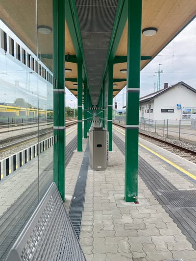 The image depicts a modern train station platform in Wulkaprodersdorf/Vulkaprodrštof, Austria. The platform features a covered waiting area supported by vibrant green pillars, with a wooden ceiling overhead. A glass barrier runs along the edge of the platform, reflecting a yellow and green train on the adjacent tracks. The platform is paved with interlocking gray concrete blocks, with tactile paving and a yellow line indicating the platform edge. A metal trash bin is visible, and a silver bench is partially in the foreground. In the background, a white building with a pitched roof, possibly a station building or related facility, is situated behind a metal fence. Overhead power lines and antennas are also visible. The sky is overcast.