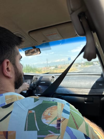 The image shows the interior of a car, from the perspective of the passenger. A man with dark hair and a beard is driving, wearing a colorful patterned shirt. His right hand is on the steering wheel. A black seatbelt is visible, diagonally across the frame. The dashboard of the car is visible, including the instrument cluster. Outside the car, through the windshield, a road is visible with trees and fields on either side, under a blue sky with some clouds. The road appears to be in a rural or semi-rural setting, consistent with the outskirts of Proshyan, Armenia.
