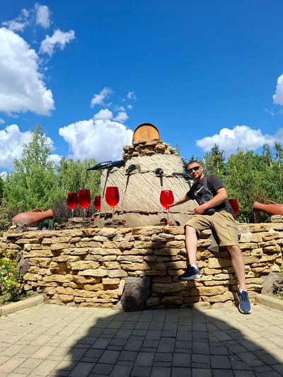 In Milestii Mici, Moldova, a man wearing sunglasses, a black t-shirt, and tan shorts sits on a stone wall in front of a fountain. The fountain is designed to resemble a large wine barrel and features several large red wine glasses, some appearing to overflow, and terracotta amphorae. A green wine bottle is positioned at the top of the fountain, with liquid appearing to pour from it into the glasses. The background consists of lush green trees and a bright blue sky with white clouds. The ground in front of the wall is paved with interlocking gray bricks, and a distinct shadow is cast across it.