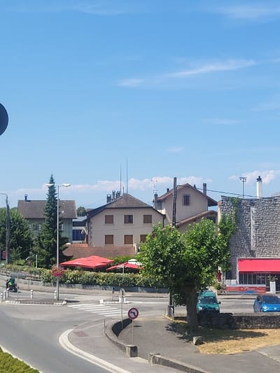 A sideways view shows a European town scene on a sunny day. Buildings of varying heights are aligned along a road with a 'no left turn' sign. Trees and greenery are interspersed between the buildings, and parked cars are visible. The sky is blue with some clouds.