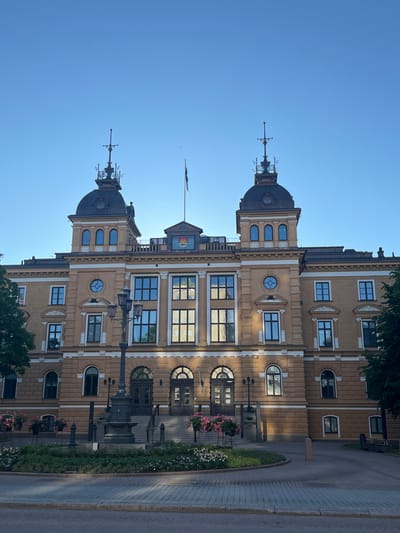 The image shows the Oulu City Hall, a prominent landmark in Oulu, Finland. The building is made of light-colored stone and features a symmetrical facade with two prominent towers topped with decorative spires. The main entrance is accessed by a grand staircase, flanked by ornate lampposts. Several windows, some arched and some rectangular, are visible on the facade, reflecting the clear blue sky. Flower boxes with pink and white flowers are placed near the entrance and along the surrounding pathways. The foreground shows a paved area with a circular garden in the center, also adorned with flowers and greenery. The sky is a deep blue, indicating it is daytime, likely late afternoon or early evening, given the soft lighting. There are no people visible in the image.