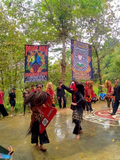 An outdoor cultural performance is captured in Tamansari, Indonesia. The scene depicts a group of individuals engaged in a traditional dance or martial arts demonstration on a wet concrete surface under the canopy of green trees. Two large banners are prominently displayed in the background, suspended between trees: the left banner features "PAGAR NUSA" text and a horned figure over a globe, while the right banner displays "PERSAUDARAAN SETIA HATI TERATE" with a heart and lotus symbol.
In the foreground, two main performers are central. The individual on the left, facing right, holds a red, shield-like object inscribed with "RAHAYU" and "SDM." This performer wears dark trousers, a patterned sarong, and a headpiece with long, dark strands, with dark makeup visible around the eyes. The individual on the right, facing left, wears a red mask with a pronounced nose, dark long hair/wig, a dark jacket, patterned sarong, and dark trousers. Their left arm is raised, and the right arm is bent. Both lead performers are barefoot.
In the mid-ground, other performers or participants are visible, some wearing light-colored, shaggy wigs or headpieces, and similar attire. To the far right, one individual is kneeling, while others stand. To the left, several young men in casual attire (t-shirts, shorts/pants) observe the event. The concrete ground is wet, showing reflections, and a circular red and white pattern is visible on the surface to the right of the performers. A hand holding a smartphone is partially visible in the bottom-left corner of the frame.