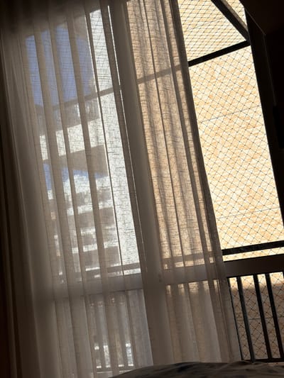 The image shows a view from inside a building, looking up through drawn blinds towards a window. Outside the window, a netting-like structure is visible, filtering the sunlight. The scene appears to be located in Brasília, Brazil, suggested by the architectural style of the unseen buildings outside the window, implying urban context. The blinds partially obscure the view, casting shadows and creating a striped effect on the visible exterior. No people or subjects are directly visible.