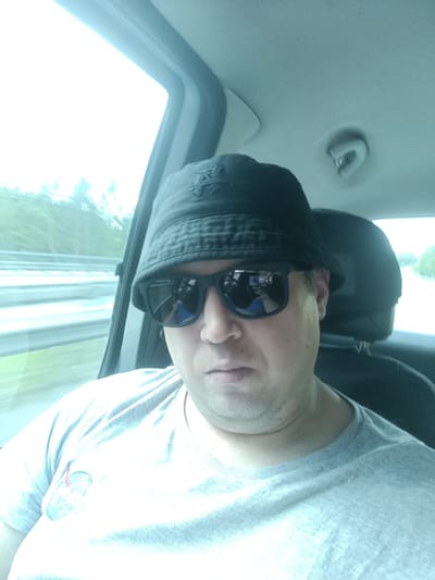 An adult individual with fair skin is depicted in the front passenger seat of a vehicle, traveling in or near Hvězdonice, Czechia. The person is wearing a dark-colored bucket hat, which features an embroidered monogram resembling interlocking "AL" letters on the front. Dark-rimmed, mirrored sunglasses cover the eyes, reflecting parts of a blue garment and interior car elements. The individual is dressed in a light grey, short-sleeved, crew-neck t-shirt, with an indistinct circular patch or sticker affixed to the upper left chest area. A noticeable double chin is present.
The vehicle's interior has light-colored ceiling and pillar panels. A grey overhead grab handle is visible above the passenger's right side. Through the blurred windows, a multi-lane road with guardrails and distant green foliage is discernible, indicating the vehicle is in motion.
