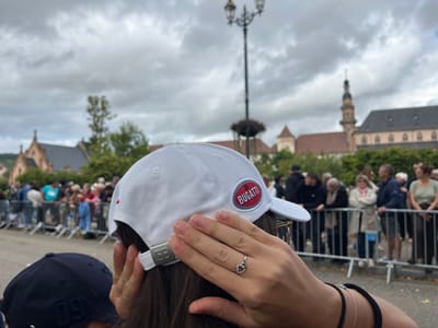 The image displays an outdoor public gathering in Molsheim, France, captured from an over-the-shoulder perspective. In the immediate foreground, the back of a person's head with dark hair is visible, wearing a white baseball cap. The cap features a circular red patch on its left side with "BUGATTI" written in white. The cap's rear adjustable strap has a silver-colored rectangular buckle embossed with the "BB" logo. The individual's right hand is raised towards their head, with fingers lightly touching the temple area. A silver-colored ring with a dark, circular design is worn on the ring finger, and a thin bracelet along with a thicker dark band are on the wrist. Partially visible in the lower-left foreground is the back of another person's head wearing a dark blue baseball cap with "09" stitched in white.

In the midground, a crowd of numerous individuals stands behind silver-colored temporary metal crowd control barriers, all facing in the same direction, observing an unseen event. The ground is a paved surface.

The background reveals architectural features characteristic of Molsheim. Prominently positioned in the central-right background is the Église des Jésuites (Jesuit Church), identifiable by its tall, multi-tiered ornate spire, dark roof, and light-colored stone facade. To the left, another reddish-brown stone building with a pointed gable and arched windows is visible amidst green foliage. The sky is overcast with a uniform layer of grey clouds.