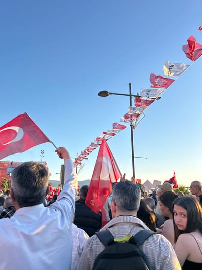 The image shows a crowd of people gathered in İzmir, Türkiye (38.437, 27.141), with a line of Turkish flags strung across a streetlamp. The people are looking towards something out of frame, and some are holding Turkish flags. One man is wearing a white shirt and raising his arm, holding a Turkish flag. He is in the foreground, while a man with a backpack is directly behind him. There is a young woman with long dark hair standing at the edge of the crowd, looking towards the camera. The sky is blue and the sun is shining.