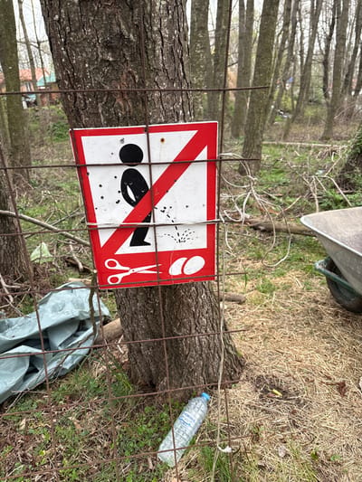 The scene, located in a partially cleared, forested area in Smolari, North Macedonia, focuses on a prominent warning sign attached to a tree trunk. The sign is a rectangular white panel with a red border and a thick red diagonal line indicating prohibition. It features a black stick figure in profile, depicted urinating, with visible splash marks. Below this primary image, a pair of white scissors is positioned on the left, and two white oval shapes, resembling eggs, are on the right, implying a severe deterrent against urination in this area.
The sign is secured to a rusty, brown wire mesh fence, which in turn is affixed to a mature tree trunk with rough, dark brown bark. In the immediate foreground to the left of the tree, a crumpled grey tarp rests on the ground. A clear plastic water bottle, labeled "ZVORSKA," lies horizontally on the sparse ground cover, which comprises green shoots and dry, yellowish grass or straw. To the right, a silver-colored wheelbarrow with a green wheel is partially visible. The background reveals a dense stand of deciduous trees with bare branches, suggesting late winter or early spring conditions. Through the distant trees, parts of residential structures with colored roofs are discernible. The sky appears overcast. No individuals are present within the frame.