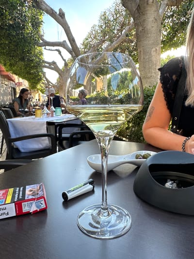 Here is a description of the image:
The image depicts a table at an outdoor restaurant in Rojales, Spain. On the table, there is a glass of white wine, a small dish containing olives, a lighter, and a pack of cigars with a warning label in Spanish about the risks of smoking. A person is visible in the background, and a woman's arm and a black top are in the upper part of the frame. The scene is bathed in sunlight, and the reflections in the wine glass show a lively outdoor dining atmosphere.