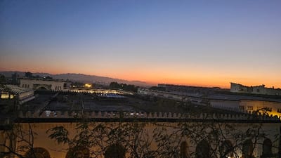 A cityscape under a vibrant dawn sky, transitioning from deep blue to orange at the horizon. Buildings with decorative facades and rooftop lights are visible, punctuated by tree branches in the foreground. Distant hills add depth. The scene evokes a tranquil yet urban atmosphere.