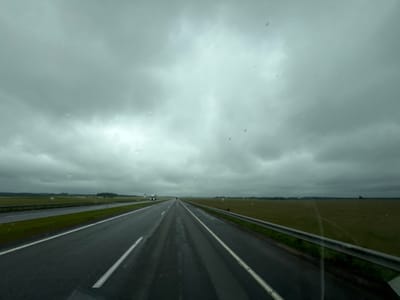 The image shows a view from a vehicle traveling on a wet, multi-lane highway under a cloudy, overcast sky. The road surface is dark and appears to be reflecting the dim light. White lines mark the lanes, with one lane to the left and two lanes visible to the right, separated by a grassy median. A metal guardrail runs along the right side of the highway. On the left, beyond the highway, a grassy field stretches to the horizon. On the right, beyond the guardrail, a similar grassy field is visible. In the distance, a few vehicles can be seen on the highway. The sky is filled with thick, grey clouds, suggesting a rainy or humid day. The image is captured as if through a windshield, with a few small spots that might be water droplets or smudges on the glass. This scene is characteristic of a highway environment in the countryside, potentially near Талачынскі сельскі Савет, Belarus, during inclement weather.
