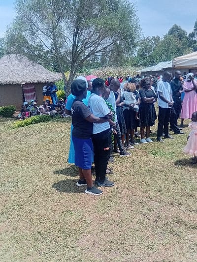A large gathering of individuals is observed outdoors in Butali Chegulo ward, Kenya. The scene unfolds on a dry, grassy expanse under a bright sky. In the foreground, a group of approximately ten adults and adolescents stands in a loosely formed line, facing generally towards the right of the frame. One woman in a black t-shirt and blue skirt stands with her back partially to the camera, next to a person in a white t-shirt and black pants. Various other attendees are dressed in a mix of casual attire, including black, white, blue, and pink garments; some are holding infants. To the far left, a traditional round hut with a thatched roof and light brown walls features a patterned fabric hanging at its entrance. A large, partially defoliated tree stands behind this hut. In the mid-ground and background, numerous other people are present, with some sitting on the ground near the hut. Further to the right in the background, a white tent canopy is partially visible. The area is bordered by dense green foliage and trees in the distance. Sunlight indicates clear weather, casting short shadows.