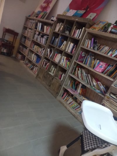 This interior scene, located in Drocourt, France, depicts a room furnished with multiple wooden bookshelves. Four distinct light-toned wooden bookshelves, densely packed with books of varying sizes and spine colors, extend along a light-colored wall from the left mid-ground to the right foreground. Among the visible titles are "Atlas" and "Barbie." Above the central bookshelves, two large rectangular art pieces lean against the wall; one displays a pink floral design, and the other features a red abstract shape. In the distant left, a small, dark-stained wooden table with intricate legs is partially visible. The immediate right foreground contains a white high chair, equipped with a white tray and a black-and-white checkered fabric seat cushion. The floor is covered with light grey rectangular tiles. The scene is devoid of human presence or activity.