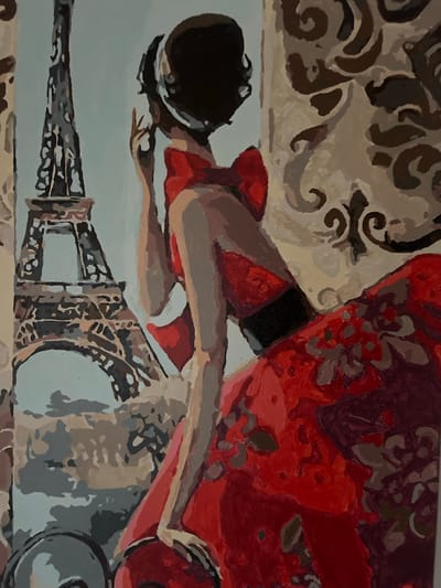 The image is a painting of a woman in a red dress standing in front of the Eiffel Tower. The woman is looking over her shoulder with her hand near her face. The painting is done in a style that resembles paint-by-numbers, with distinct blocks of color and visible brushstrokes. The background includes a stylized representation of the Eiffel Tower and a patterned wallpaper. The lighting appears to be soft and diffused. The specific location in Vorniceni, Moldova is not discernible from the image itself.