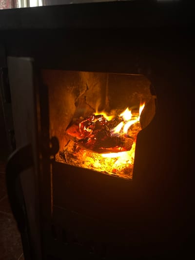 The image displays an interior scene in Lesidren, Bulgaria, centered on a dark-colored wood-burning stove or fireplace. Inside the firebox, wood logs are actively burning, producing vibrant orange and yellow flames, alongside a bed of glowing red embers and grey ash. The stove's access door on the left is partially open, exposing a dark, curved handle. The fire is the dominant light source, illuminating the firebox and casting a warm, dim glow on the surrounding environment, which remains largely dark. Faint reddish-brown patterned floor tiles are discernible in the lower-left portion of the frame, adjacent to the stove. No individuals are visible.