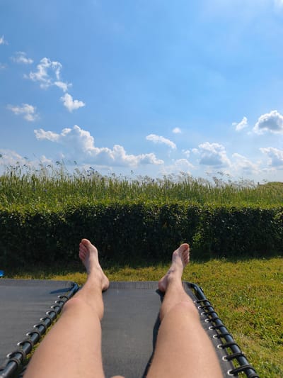 The image depicts a person's legs and feet resting on a dark-colored lounger. The lounger is positioned on a green lawn. Beyond the lawn is a thick hedge, and behind the hedge is a field of tall green grass and some plants with blue flowers. The sky above is a clear blue with scattered white clouds. The scene appears to be outdoors on a sunny day, possibly in a residential area or park in Kyviškės, Lithuania.
