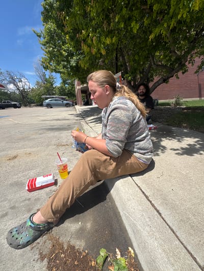 The image captures a scene on a sidewalk in Lafayette, Indiana, United States. A young woman is hunched over, looking at a bag of chips in her hands. She is wearing a grey, patterned hooded sweatshirt, brown pants, and patterned Crocs. Scattered on the pavement next to her are a Big Gulp cup and a box from Raising Cane's Chicken Fingers. In the background, a man with long hair and a beard is sitting under a tree. Cars and buildings are visible further in the background. The scene appears to be outdoors, with a clear blue sky visible through the trees.