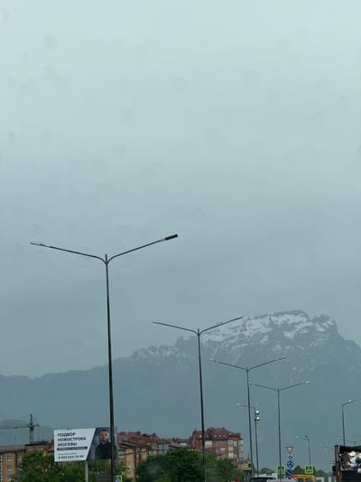 The image depicts a cloudy, overcast sky over a city with snow-capped mountains in the background. In the foreground, several streetlights are visible, along with a billboard advertising real estate. The billboard features an image of a man and text in Russian, indicating services related to new buildings in Moscow. Residential buildings and trees are also present, as well as traffic signs and a construction crane. The overall scene suggests an urban environment with a backdrop of natural mountainous terrain, under a muted and possibly rainy weather condition.