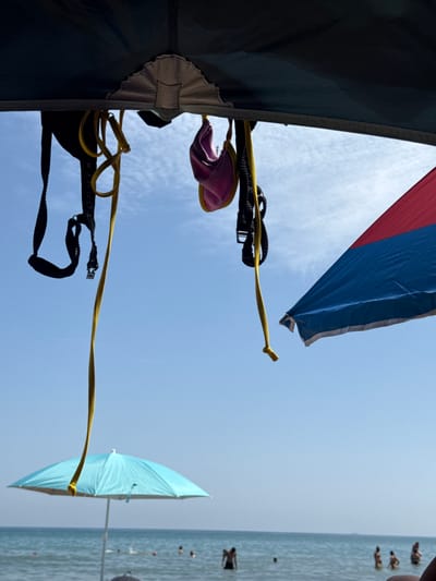 A low-angle, close-up shot captures the underside of a dark-colored tent or canopy against a clear, bright blue sky dotted with wispy white clouds. Several items are hanging from the canopy: a black strap with a silver buckle, a yellow rope twisted into a loop, and a small, pink bikini top. To the right, a portion of a red and blue beach umbrella is visible, angled upwards. In the background, a bright turquoise beach umbrella is opened and positioned over the sea. The ocean is calm, with faint ripples on the surface. Several people are visible wading or swimming in the shallow water. The scene appears to be set at a beach, likely in Gavà, Spain, on a sunny day.