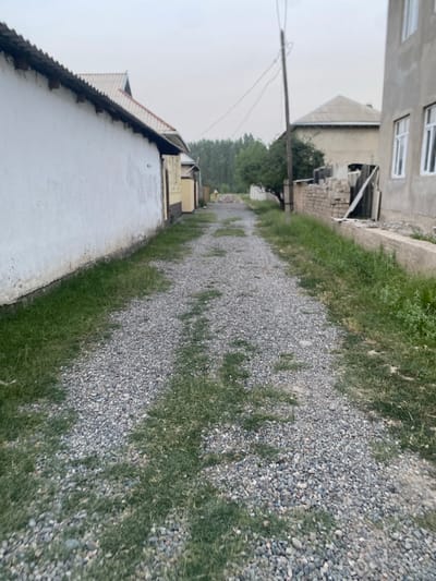 The image depicts a narrow, unpaved pathway bordered by grass and gravel, winding through a residential area. On the left, a long, white-walled building with a corrugated roof stretches along the path. Further back, a two-story building with multiple windows is visible, alongside a pile of building materials, suggesting construction or ongoing repairs. The pathway leads towards a cluster of trees in the distance, indicating a rural or semi-urban setting. The sky is overcast, suggesting a cloudy day, and the overall lighting is dim, hinting at either early morning or late afternoon. There is no discernible activity or people in the image, giving it a quiet and still atmosphere. The location is identified as Боғистон, Tajikistan.