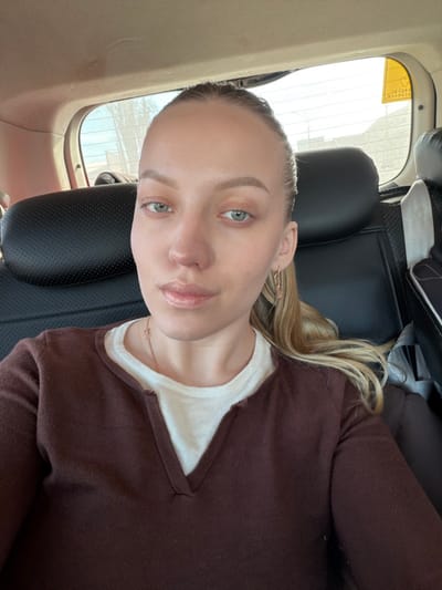 The image depicts a female individual seated in the rear passenger compartment of a vehicle, facing the camera. Her head is centered, and her gaze is directed forward. She has light skin, light-colored eyes, and her light-brown hair is pulled back, with some strands visible over her right shoulder. She is wearing a dark brown long-sleeved V-neck top over a light-colored V-neck undershirt. A delicate gold-colored chain necklace with a small pendant is visible on her neck, and a gold-colored elongated hoop earring is present in her right ear. The interior of the vehicle shows black headrests and a light-colored ceiling. Through the rear window, an indistinct outdoor environment with bright sky is visible. A yellow rectangular sticker with black Cyrillic text, consistent with "РЕБЕНОК" (Child), is affixed to the upper right corner of the rear window. The specific location within the city of Zavidovo, Russia, cannot be confirmed from the visual evidence.