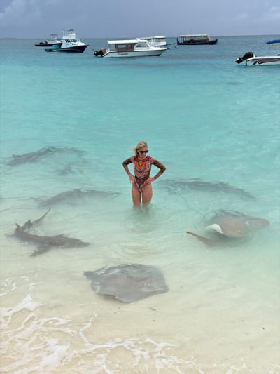 A medium shot shows a blonde woman standing in the shallow, turquoise waters of Fulidhoo, Maldives. She is wearing a multicolored, patterned swimsuit and sunglasses. Around her, several stingrays swim. To the left of the frame, boats are anchored. The water transitions to white sand and small waves on the right. The sky is overcast on the left side of the frame.