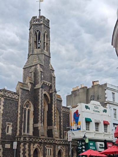 The image depicts a street scene in what appears to be a town or city in the United Kingdom, characterized by a combination of historical and modern elements. The scene is dominated by a large stone building with a prominent tower, likely a church, displaying Gothic-style architecture with arched windows and intricate stonework. Adjacent to the church, a white building houses a "Pizza Pilgrims" restaurant, adorned with green and red awnings and a large mural of a figure resembling Spider-Man. Red umbrellas suggest outdoor seating for the restaurant. Signage for a "Pedestrian and Cycle Zone" is visible near the pizza restaurant. The sky is overcast with mammatus clouds. The OCR mentions phrases visible on the image: "PIZZA PILGRIMS", "PEDESTRIAN and CYCLE ZONE", "HOT PIZZA", "COLD PIZZA". The precise location within Reigate and Banstead cannot be determined solely from the image.