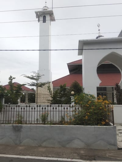 The image depicts a white mosque with a tall, slender minaret in Lancok, Indonesia. The mosque features a red-tiled roof and a distinctive arched entryway. The minaret is topped with a crescent moon symbol. In front of the mosque, a metal fence encloses a garden with flowering bushes and trees. The scene is captured on an overcast day, with power lines visible in the foreground, obscuring parts of the sky and structures. The ground in front of the garden is a paved walkway next to a road.