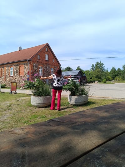 A woman with long brown hair, wearing a black and white polka dot t-shirt and bright pink pants, stands with her back to the camera in an outdoor setting. Her arms are outstretched to her sides. She is positioned in front of a large, round planter filled with green foliage and some small purple flowers. Behind her, to the left, is a stone and brick building with a red tile roof and several windows. A sign with an orange and yellow graphic is visible to the left of the building. To the right of the building, there is a paved area, likely a parking lot, with several vehicles parked. Trees and vegetation are present in the background. In the foreground, a dark wooden table surface is visible, partially obscuring the lower portion of the scene. The sky above is blue with scattered clouds. The overall impression is a sunny day in a semi-rural or coastal location, possibly near a historical building. The image captures a moment of relaxed enjoyment or appreciation of the surroundings. The building's architecture suggests it might be a former industrial or agricultural structure. The presence of parked vehicles and the paved area indicate public access. The scene appears to be located in Pāvilosta, Latvia, known for its coastal scenery and historic buildings.