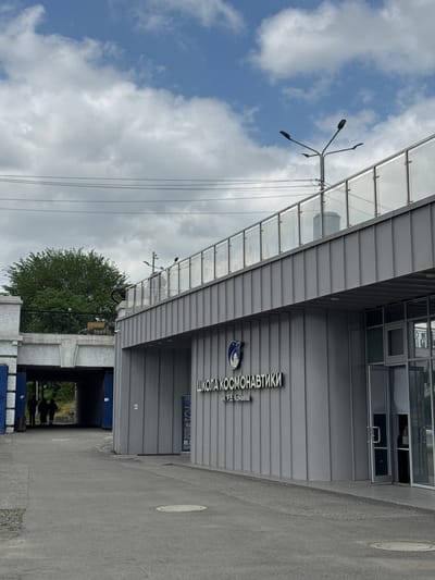 The image captures the exterior of the "School of Cosmonautics" named after P.B. Konaeva in Zamankul, Russia. The building is characterized by its gray, corrugated facade and glass-paneled awning supported by metal frames. Streetlights extend from the awning towards a partly cloudy sky. A doorway, leading to an enclosed passage, is visible under the building. A group of figures are silhouetted within this passage, and a section of trees lies beyond it. The ground surface is a gray pavement. The sky occupies the upper-left portion of the frame, featuring scattered clouds and patches of blue sky.