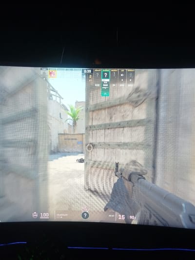 The image displays a digital screen, likely a monitor, showing a first-person perspective from a tactical video game. The environment depicted within the game is a virtual desert setting featuring Middle Eastern-style architecture, including sand-colored stucco buildings and weathered wooden doors, and is not a real-world location in Balykchi, Uzbekistan. The foreground shows an open area beyond two large, partially open wooden doors with metal bands and a ring handle on the right door. A palm tree is visible in the background against a bright sky. A small, dark object rests on the ground near the left door.
In the lower right quadrant, a player character's right arm, covered in a textured, possibly camouflage, sleeve, holds an AK-47 assault rifle, pointed forward. The rifle's front sight is clearly visible.
Overlaying the game environment are user interface elements:
* A timer "0:39" is centered at the top.
* Player statistics "4 | 2" and an avatar for "PERBUFF" are at the top left.
* Other player statuses and scores, including a highlighted green box with "101" and "$5000," are displayed at the top right.
* The bottom left shows "100" next to a shield icon, indicating health or armor, and the player name "clicKilleR01".
* The bottom right displays the weapon "AK-47" with ammunition counts "15 | 90."
The photograph captures a screen door effect, indicating the pixel grid of the display. The physical bezel of the monitor, dark in color, frames the top and bottom of the active display area, with a faint blue light visible at the very bottom edge.