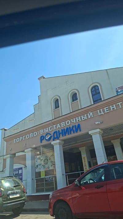 The image shows the facade of the "Rodniki" trade and exhibition center in Dzyarzhynsk, Belarus. The building is light-colored with arched windows. Above the entrance, large blue lettering spells out "РОДНИКИ" (Rodniki) along with the text "ТОРГОВО-ВЫСТАВОЧНЫЙ ЦЕНТ" (Trade and Exhibition Center). Several cars, including a green Dacia Duster and a red car, are parked in front of the building. The sky is clear and blue.