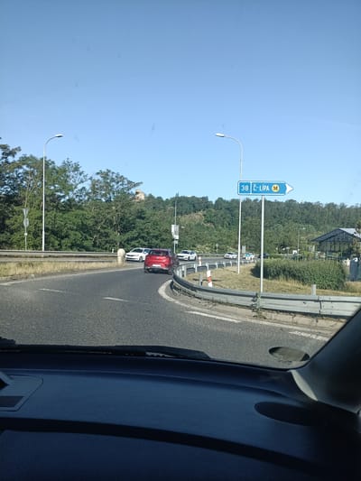 The image shows a view from a vehicle's dashboard, looking out at a road with a slight curve to the right. A blue road sign indicates "38 Č-LÍPA M" to the right, suggesting this is a route towards Česká Lípa. Several cars are visible on the road, including a red car in the foreground and white cars further ahead. Trees and vegetation line the sides of the road, with a hill in the background. The sky is clear and blue. The scene appears to be in Mladá Boleslav, Czechia, given the road sign.